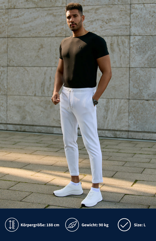 Model trägt ein schwarzes Slim Fit T-Shirt mit einer weißen Hose und steht vor einer grauen Wand.